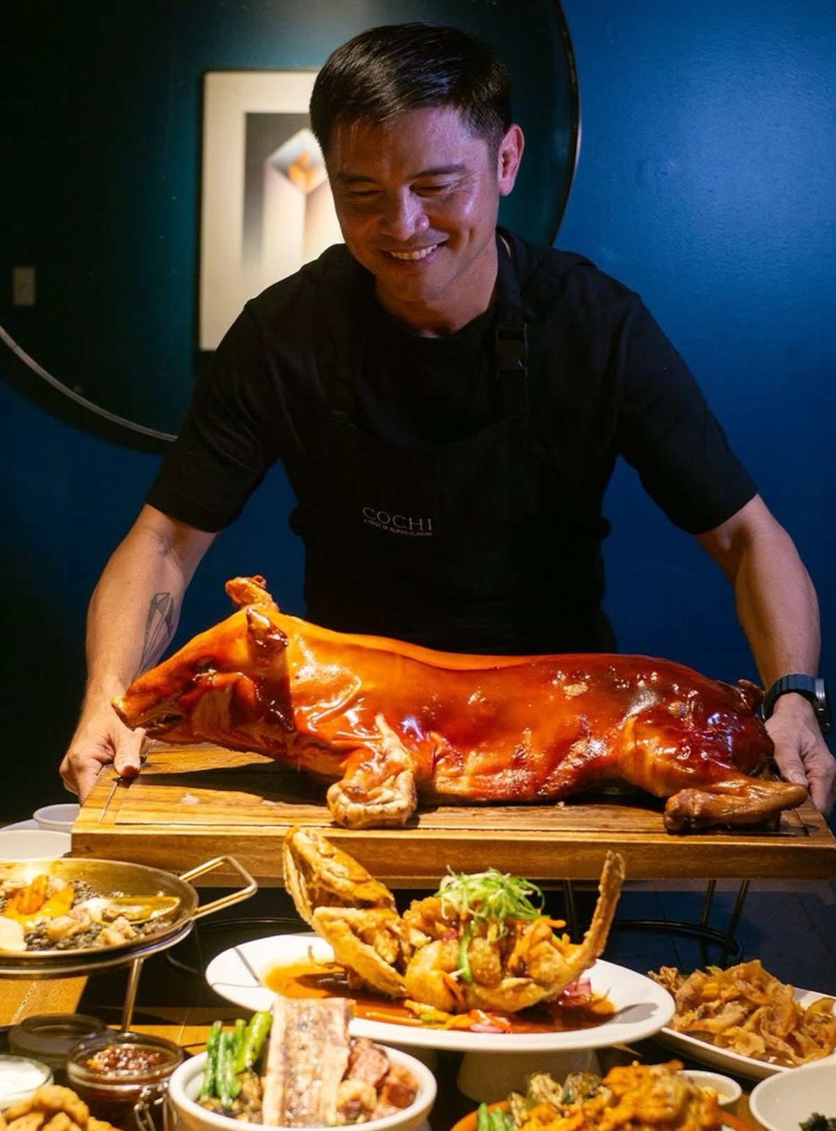 A restaurant host presenting a whole cochinillo above a table of Filipino dishes